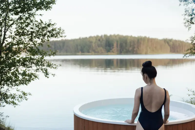 A woman in a swimsuit relaxes in an outdoor hot tub by a serene lake surrounded by trees