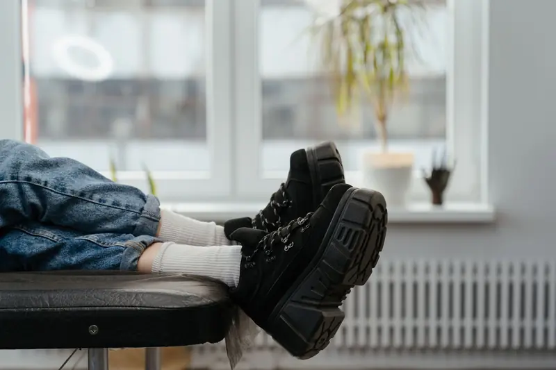 Person in a tattoo studio wearing blue jeans and black boots, relaxing by the window