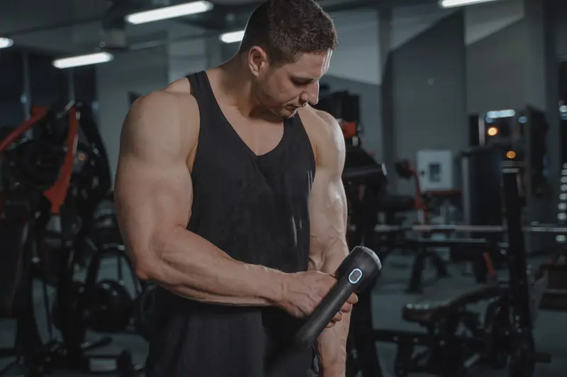 A muscular man uses a massage gun in an indoor gym, focusing on recovery