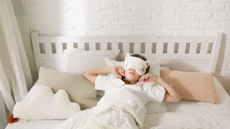 A young girl resting in bed with a cute sleep mask surrounded by cozy pillows