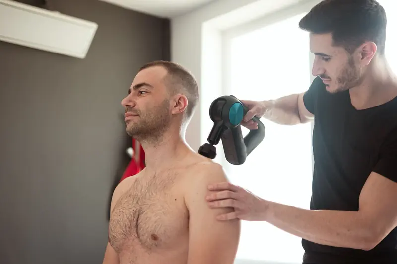 A physiotherapist provides shoulder massage therapy with a massage gun in a clinic