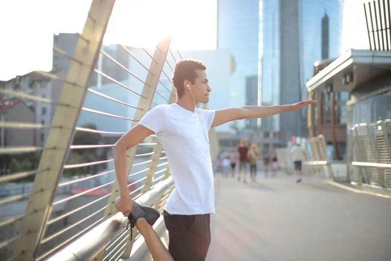 Low angle serious concentrated African American man in earphones wearing white shirt and black shorts stretching legs and listening to music with pleasure on st